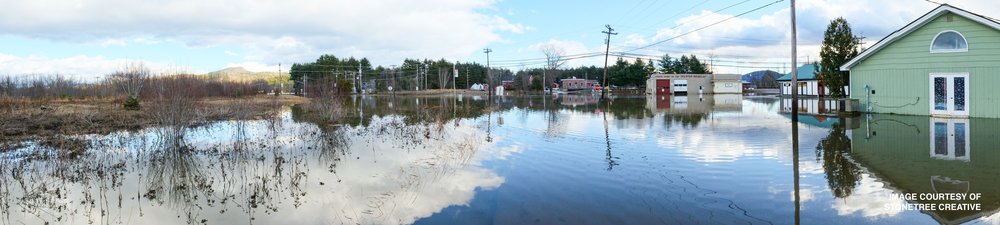 Photograph of flooded buildings with water flowing from the buildings into a river with mountains in the background